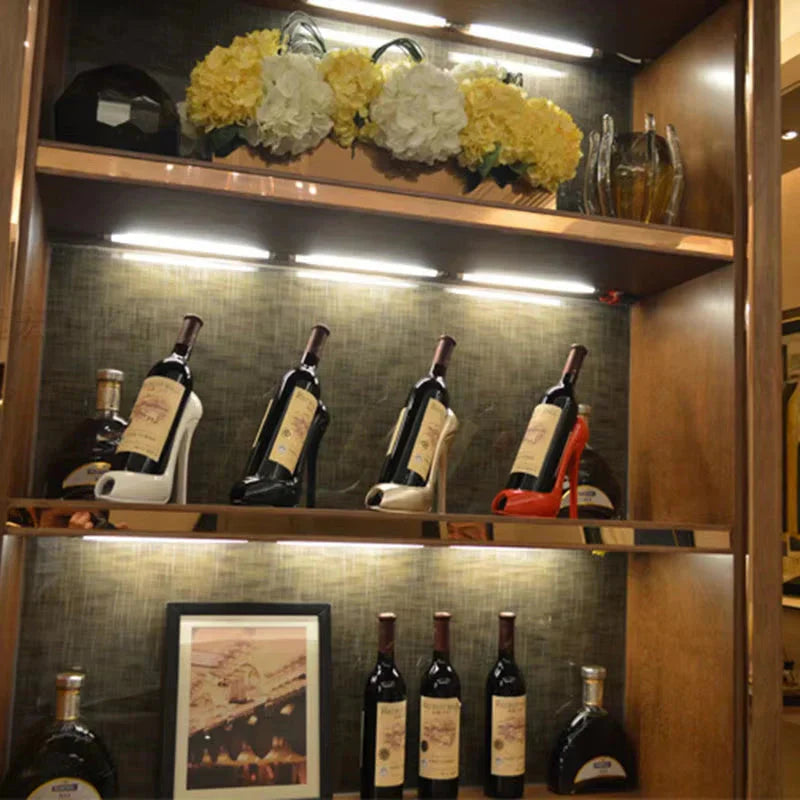 Wine bottles displayed on high heel holders in a lit wooden shelf with floral decor